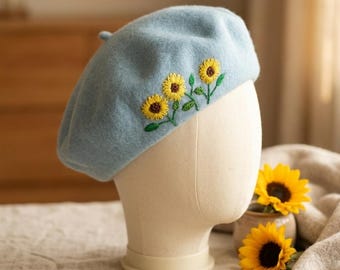 Puede incluir: Boina azul claro con bordados de girasoles amarillos y hojas verdes. La boina se exhibe en una cabeza de maniquí. Un accesorio de moda.