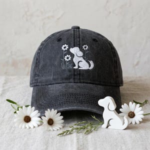 Op de afbeelding: Donkergrijze baseballcap met een wit geborduurd ontwerp van een hond en bloemen. De cap wordt getoond met witte madeliefjes en een klein wit hondenbeeldje.