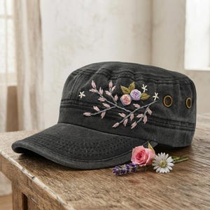 Op de afbeelding: Een antracietgrijze pet in militaire stijl met geborduurde bloemendetails aan de zijkant. Het borduurwerk bevat roze en paarse rozen met groene bladeren en kleine witte stervormige bloemen. Twee koperkleurige metalen oogjes zijn zichtbaar.