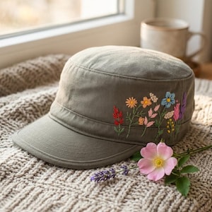 May include: An olive green cadet cap featuring detailed floral embroidery on the side. The embroidery depicts a variety of small wildflowers in shades of red, orange, pink, blue, and yellow. The cap has a flat top and a short, stiff brim.