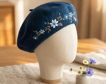 Può includere: Un basco blu navy con fiori bianchi e azzurri ricamati e steli color oro. Il basco è esposto su una testa di manichino. Un piccolo vaso con fiori è su un tavolo sullo sfondo.