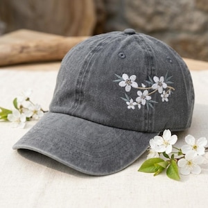 May include: A gray baseball cap with a curved brim, featuring an embroidered floral design of white blossoms and green leaves on the side. The cap is made of a soft, textured fabric.