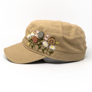 以下が含まれることがあります： ベージュ色のミリタリースタイルのキャップ、花の刺繍入り。 フラットなつばと構造化されたクラウンが特徴です。 花の刺繍には、白、ピンク、茶色、青の色合いのデイジーやその他の花が含まれています。 耐久性のある生地で作られたキャップです。