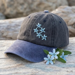 Può includere: Un cappellino da baseball grigio scuro con visiera blu e fiori ricamati azzurri. Il cappellino è realizzato in tessuto morbido e ha una visiera curva. Il cappellino è appoggiato su una superficie di legno.