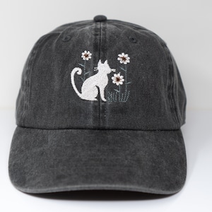 Puede incluir: Una gorra de béisbol de denim negro con un gato blanco bordado sentado en un campo de flores blancas.