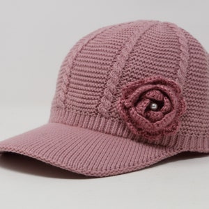 May include: A pink knitted baseball cap with a large pink crocheted flower on the top. The flower has a pearl in the center.