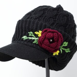 Puede incluir: Boina negra de punto con visera y una flor burdeos bordada en el lateral. La flor tiene detalles amarillos y hojas verdes.