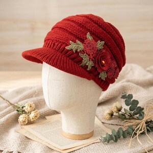 May include: A red knit hat with a small brim, adorned with a floral applique. The hat is displayed on a mannequin head. The floral design features red flowers and green leaves. The hat's texture is ribbed, and the color is a deep, rich red.