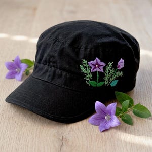 Puede incluir: Gorra negra de estilo militar con un pequeño diseño floral bordado en morado y verde. La gorra tiene una visera corta y una corona estructurada. Dos flores moradas y hojas verdes se colocan cerca de la gorra.