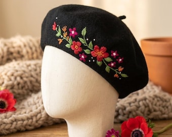 May include: A black beret with embroidered floral detailing. The embroidery features red, pink, and orange flowers with green leaves. The beret is displayed on a mannequin head, with a wooden surface in the background.