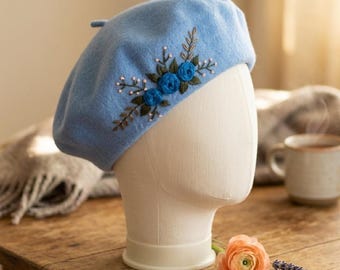 Puede incluir: Boina azul claro con rosas y hojas azules bordadas. La boina se exhibe en una cabeza de maniquí. La boina está hecha de un material suave y está decorada con pequeñas cuentas blancas. Un accesorio de moda.