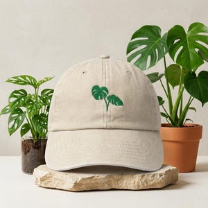 May include: A beige baseball cap with a green embroidered plant design. The cap is made of a soft fabric and features a curved brim. The design is a stylized depiction of two leaves. The cap is displayed on a stone surface, with potted plants in the background.