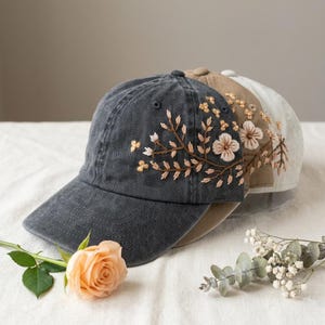 May include: Three baseball caps in shades of gray, tan, and off-white, each adorned with embroidered floral designs. The caps are arranged in a row, with a peach rose and greenery in the foreground.