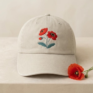 May include: A beige baseball cap with an embroidered floral design featuring two red poppies and green leaves. A single poppy flower lies next to the cap. The cap has a curved brim and a button on top.