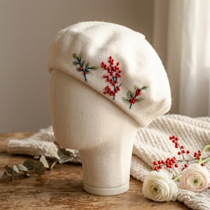 May include: An off-white felt beret featuring hand-embroidered green leaves and clusters of small red berries. The embroidery is positioned on the side of the beret, showcasing detailed stitching and vibrant red bead accents.