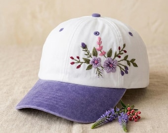 May include: A white baseball cap with a purple brim and embroidered floral design on the front panel. The embroidery features purple and pink flowers with green leaves and stems. Small purple eyelets are visible on the top of the white crown.