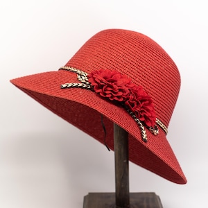 May include: A vibrant red straw hat with a wide brim, adorned with a red flower and a braided band with gold and black detailing. The hat is displayed on a wooden stand.