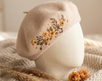 May include: A beige beret with embroidered yellow flowers and green leaves. The floral design is on the side of the hat. The beret is displayed on a mannequin head. Dried flowers and wheat stalks are in the foreground.