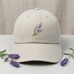 Peut inclure: Casquette de baseball beige avec une branche de lavande brodée. La lavande a des fleurs violettes et des feuilles vertes. La casquette est posée sur une surface grise claire, avec des brins de lavande.