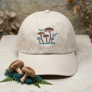 Könnte beinhalten: Beige Baseballkappe mit gesticktem Design von drei braunen Pilzen mit blauen Akzenten. Die Kappe hat einen gebogenen Schirm und einen Knopf oben. Zwei echte Pilze liegen auf grünem Moos.