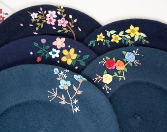 Puede incluir: Cinco boinas azules con diferentes diseños florales bordados. Las boinas están apiladas una encima de la otra, con la boina superior con un diseño floral rosa y blanco.