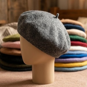 May include: A gray wool beret displayed on a mannequin head. A stack of colorful berets in various colors, including green, pink, and blue, is visible in the background. The beret is a classic, round hat.
