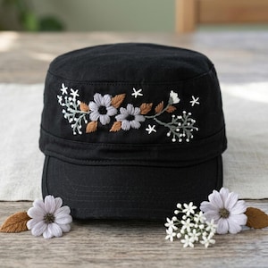 May include: Black military-style cap with embroidered floral design. The embroidery features gray, brown, and white flowers and leaves. The cap has a flat brim and is displayed on a wooden surface with decorative flowers.