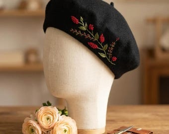 Può includere: Un basco nero con ricami di bacche rosse e foglie verdi. Il cappello è esposto su una testa di manichino. Un piccolo bouquet di fiori color pesca e un paio di forbici dorate sono su una superficie di legno.