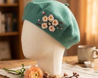 Puede incluir: Una boina verde turquesa con flores rosas y blancas bordadas. La boina se exhibe en una cabeza de maniquí. La boina es un accesorio elegante para cualquier atuendo.