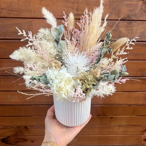 May include: A white ceramic vase with a ribbed texture holds a dried flower arrangement. The arrangement features white, pink, and beige dried flowers and grasses.