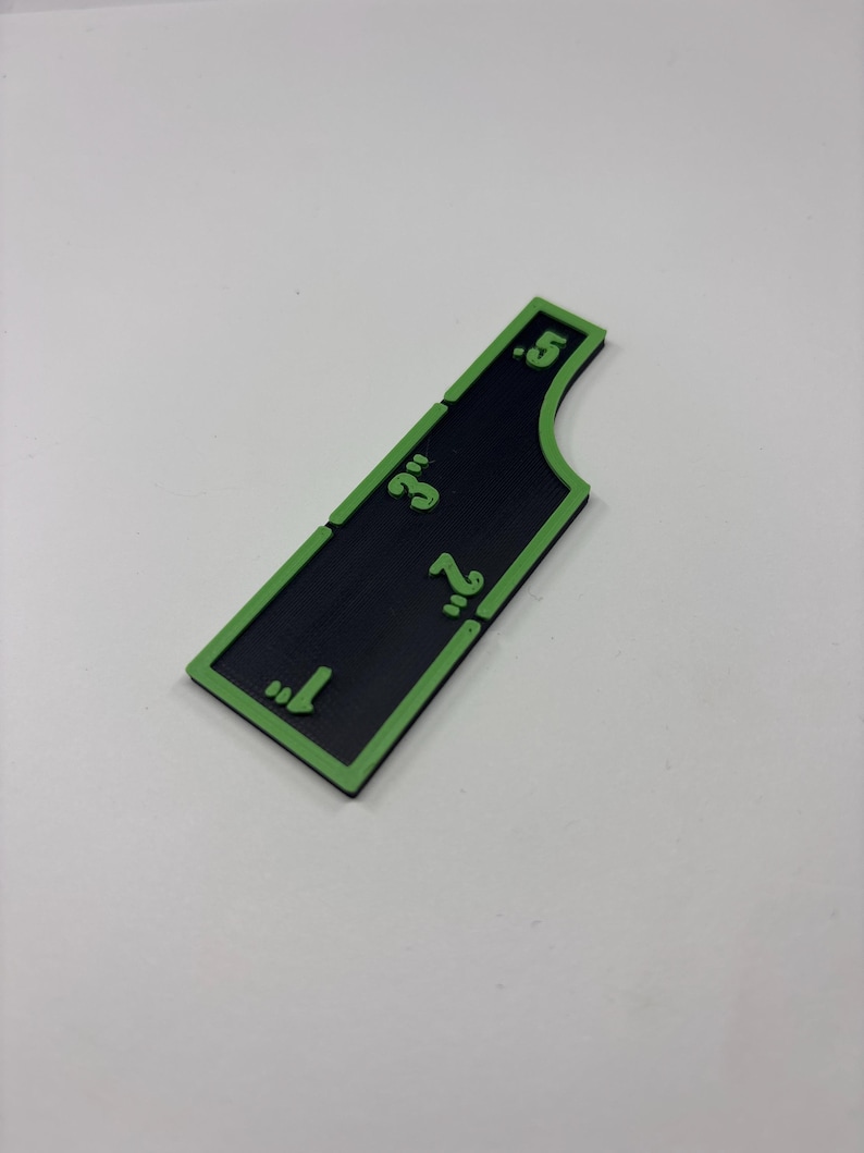 May include: A black and green measuring tool with measurements in inches. The tool has a rectangular shape with a curved edge and green markings indicating 1", 2", 3", and 0.5".