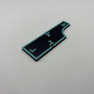 May include: A black and teal-edged ruler with measurements from 1" to 5". The ruler is rectangular with a curved edge on one side. The numbers are teal and are in inches.