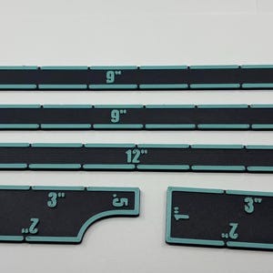 May include: A set of black rulers with teal edges and white markings. The rulers are labeled with measurements in inches, including 3-inch, 9-inch, and 12-inch lengths. The set includes two smaller, uniquely shaped rulers.
