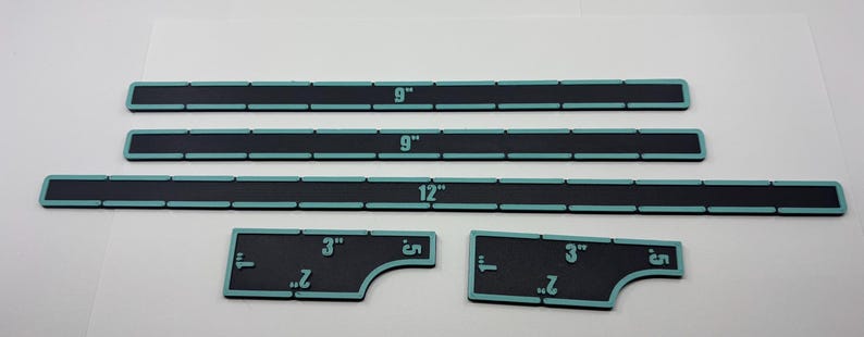 May include: A set of black rulers with teal edges and white markings. The rulers are labeled with measurements in inches, including 3-inch, 9-inch, and 12-inch lengths. The set includes two smaller, uniquely shaped rulers.