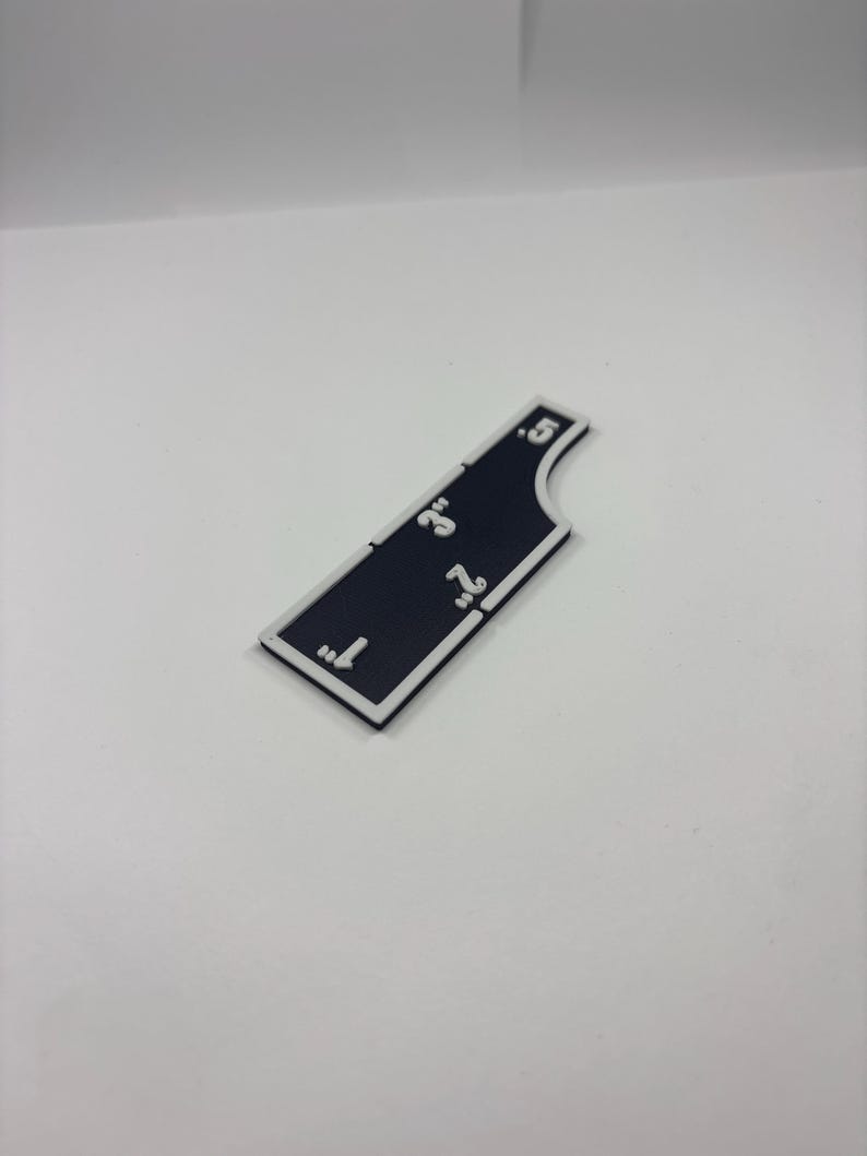 May include: A black and white measuring tool with inch markings from 1" to 5". The tool has a white border and is shaped with a curved edge. The numbers are in white.