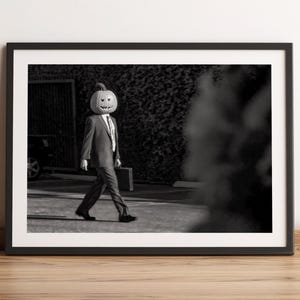 Pode incluir: Impressão emoldurada em preto e branco de uma pessoa usando uma cabeça de abóbora e um terno, andando na rua. A pessoa carrega uma pasta. A imagem é definida contra uma parede e um piso de madeira.