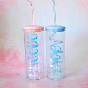 May include: Two clear plastic tumblers with matching colored lids and straws. The tumbler on the left has a pink lid and a pink straw with the word "Love" written in blue on the side. The tumbler on the right has a blue lid and a blue straw with the word "Live" written in blue on the side.