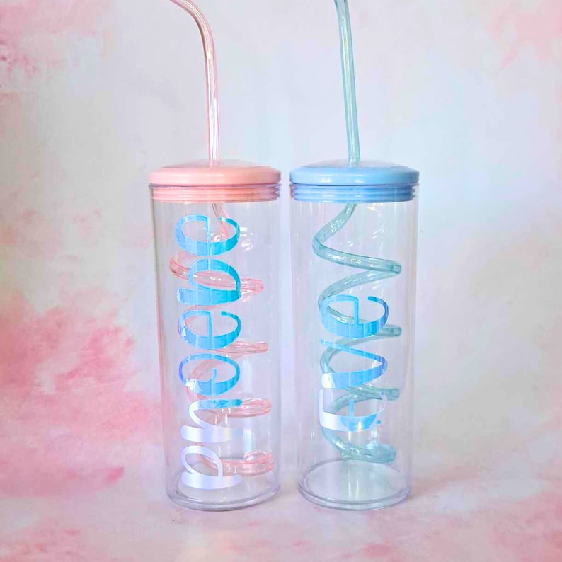 May include: Two clear plastic tumblers with matching colored lids and straws. The tumbler on the left has a pink lid and a pink straw with the word "Love" written in blue on the side. The tumbler on the right has a blue lid and a blue straw with the word "Live" written in blue on the side.