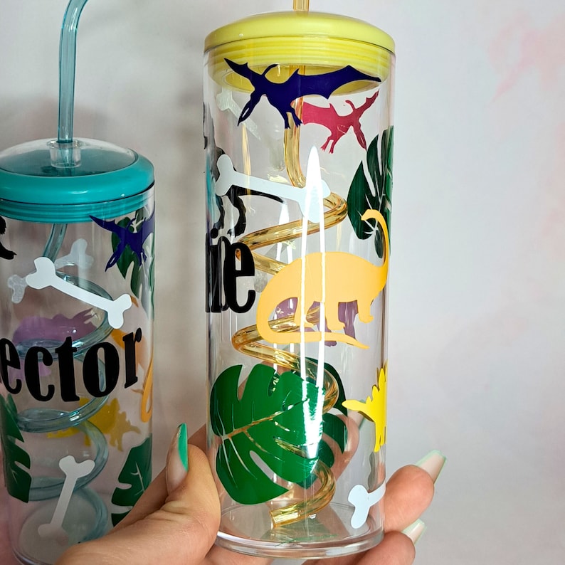 May include: Two clear plastic cups with yellow lids and straws. The cups are decorated with dinosaur silhouettes, a spiral design, and green leaves. One cup has the word "lector" written on it.