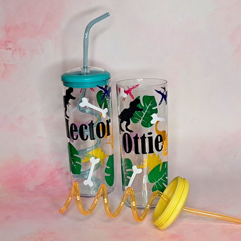 May include: Two clear plastic tumblers with lids and straws. The tumblers have dinosaur and jungle leaf designs. One tumbler says "Hector" and the other says "Ottie".