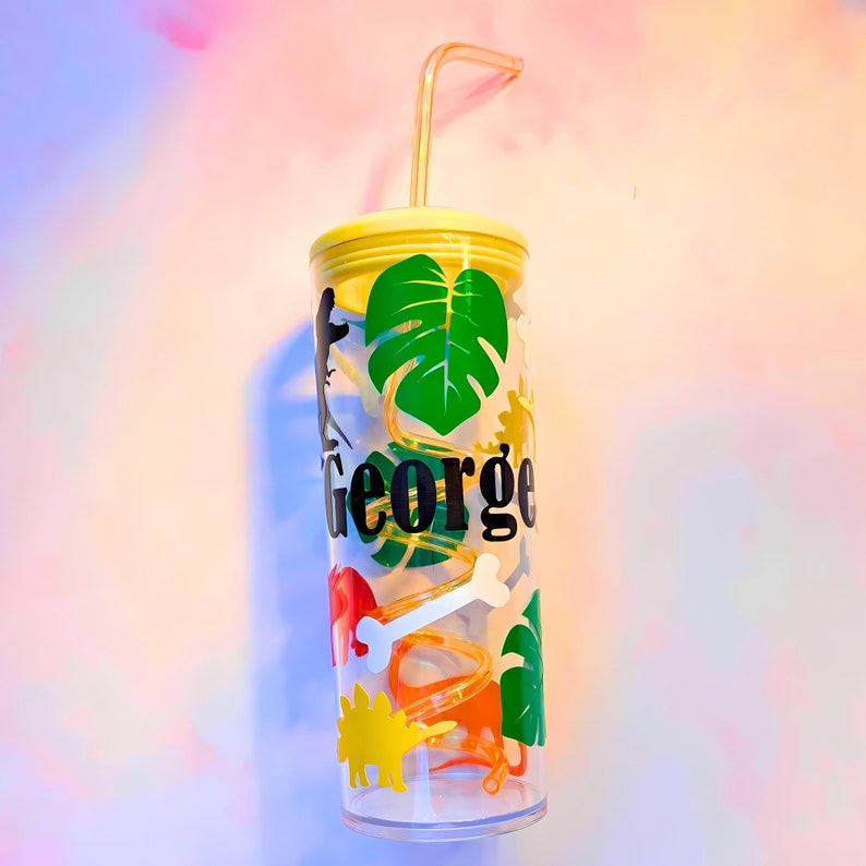 May include: A clear plastic tumbler with a yellow lid and a straw. The tumbler has a green monstera leaf, dinosaur silhouettes, and the name "George" printed on it.