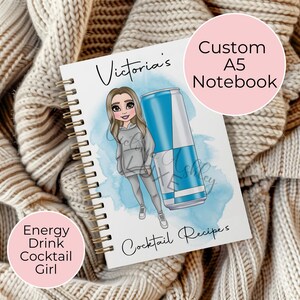 May include: A white A5 notebook with a gold spiral binding. The cover features a cartoon illustration of a girl holding a blue and white energy drink can. The text "Victoria's" is at the top of the cover and "Cocktail Recipes" is at the bottom.