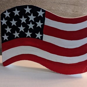May include: A wooden American flag with red and white stripes and white stars on a blue field. The flag is painted in a distressed style and is shaped like a wave.