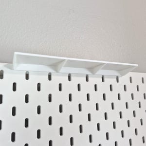 Puede incluir: Un panel perforado blanco con un patr&oacute;n repetitivo de ranuras ovaladas verticales, con un estante rectangular blanco fijado en el borde superior. El estante est&aacute; sostenido por tres peque&ntilde;os divisores verticales que crean una superficie plana.