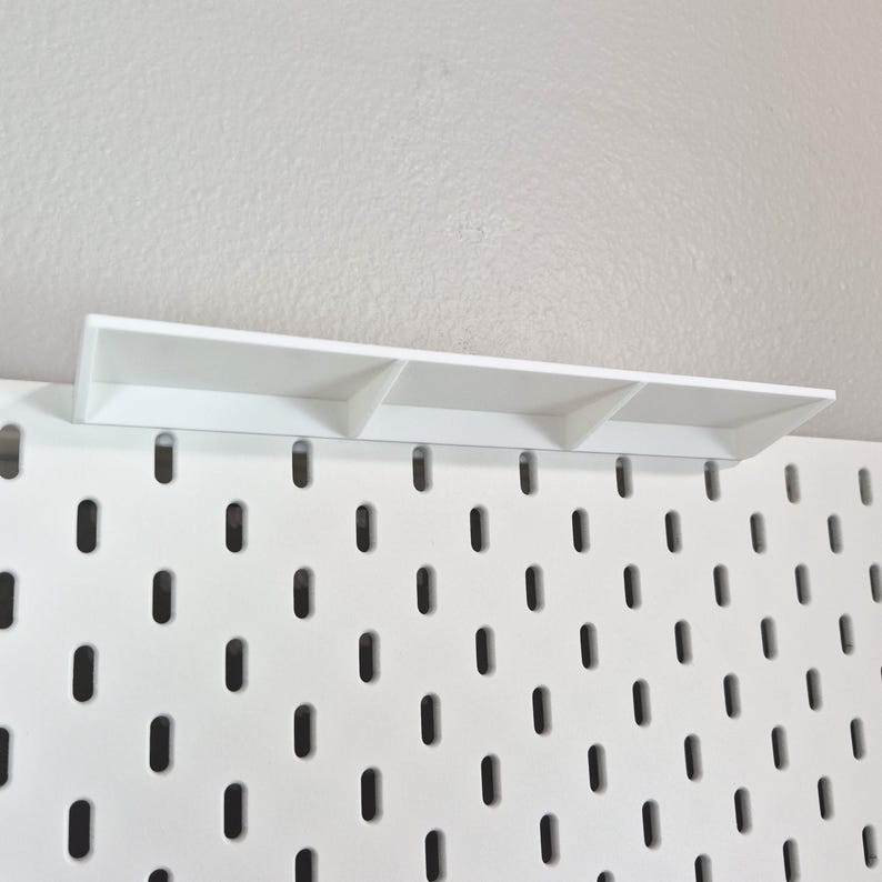 Puede incluir: Un panel perforado blanco con un patr&oacute;n repetitivo de ranuras ovaladas verticales, con un estante rectangular blanco fijado en el borde superior. El estante est&aacute; sostenido por tres peque&ntilde;os divisores verticales que crean una superficie plana.