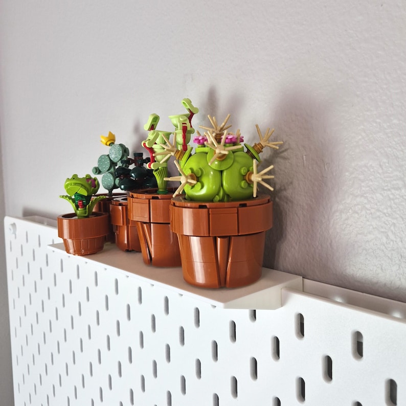 Puede incluir: Una colecci&oacute;n de cuatro peque&ntilde;as plantas en maceta construidas con bloques de construcci&oacute;n de pl&aacute;stico. Las plantas presentan varios tonos de verde, amarillo y rosa, alojadas en macetas cil&iacute;ndricas marrones sobre un estante de pared blanco.