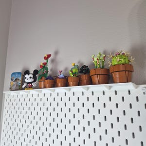 Puede incluir: Una colecci&oacute;n de peque&ntilde;os modelos de plantas en maceta construidos con ladrillos de pl&aacute;stico, que incluyen varias suculentas y una venus atrapamoscas, junto a una figura de Mickey Mouse y una lata rectangular con la etiqueta Saint-Michel.
