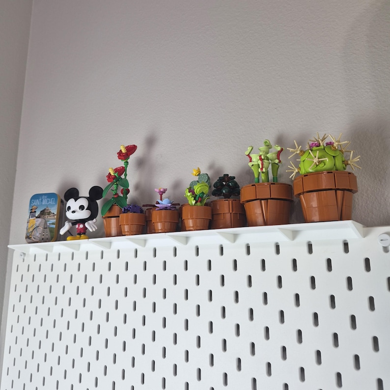 Puede incluir: Una colecci&oacute;n de peque&ntilde;os modelos de plantas en maceta construidos con ladrillos de pl&aacute;stico, que incluyen varias suculentas y una venus atrapamoscas, junto a una figura de Mickey Mouse y una lata rectangular con la etiqueta Saint-Michel.