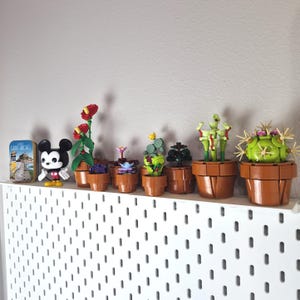 Puede incluir: Una colecci&oacute;n de peque&ntilde;as plantas de bloques de construcci&oacute;n de pl&aacute;stico en macetas marrones, incluyendo una flor roja alta, una venus atrapamoscas y un cactus redondo con espinas. Junto a ellas hay una figura de Mickey Mouse y una lata con Saint-Michel.
