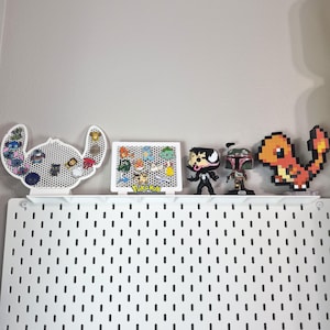 Puede incluir: Un expositor de pines blanco con forma de Stitch, un tablero rectangular blanco con la palabra Pokemon, una figura negra de Venom, una figura de Boba Fett y una figura pixelada naranja de Charmander en un estante sobre un panel perforado blanco.